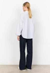 Wit en lichtblauw gestreept overhemd met een afgeronde zoom, gecombineerd met donkerblauwe flared jeans en witte sneakers, bekeken vanuit de achterkant.