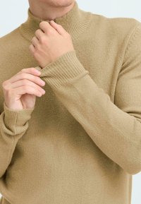 Maglione a collo alto beige a maglia con polsini e colletto a coste. Tessuto liscio con design aderente, mani che aggiustano le maniche.