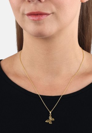 Una donna che indossa una collana con ciondolo a forma di farfalla in oro, appesa a una catena delicata, abbinata a un top nero a coste, visibile dalla mascella fino alla clavicola.