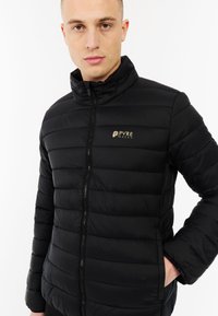 Vyras, vilkintis juodą siuvinėtą striukę su aukštu apykakle ir PVRE žalios spalvos logotipu ant krūtinės, stojantis su viena ranka kišenėje.