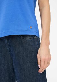 T-shirt in cotone blu con scollatura rotonda, maniche corte e un accento di logo metallico all'orlo, abbinato a jeans in denim scuri.