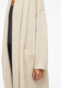 Γυναίκα που φοράει μία μακριά, κρεμ απόχρωσης κάρντιγκ με τσέπη, σε συνδυασμό με μαύρη μπλούζα και παντελόνι, με το χέρι της χαλαρό στο πλάι.