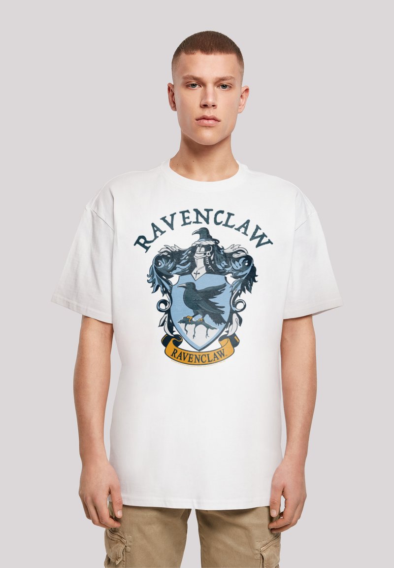 Fehér póló Ravenclaw címerrel, amelyen egy fekete holló, kék pajzs, díszítő elemek és egy sárga szalag található alul, rajta a "RAVENCLAW" felirattal.