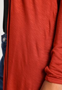 Roter strukturierter Stoff mit einem subtilen Muster liegt über einem schwarzen Material. Der Stoff hat ein sanftes Erscheinungsbild und lose, geraffte Details an den Kanten.