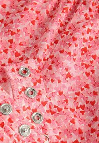 Rosa Stoff mit einem Herzmuster in roten Farbtönen, ausgestattet mit silbernen Druckknöpfen entlang einer Kante. Weiche, strukturierte Oberfläche mit einem verspielten Design.