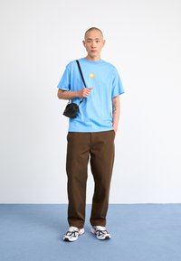 Sky blue cotton t-shirt with a small yellow logo, paired with brown pants. Black crossbody bag and white sneakers with black accents.