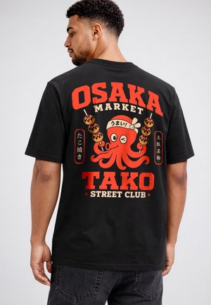 Man wearing black T-shirt with red and beige octopus holding skewers, and text "Osaka Market Tako Street Club" on the back.