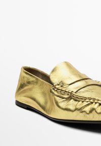 Goldene Lederloafer mit einem geflochtenen Riemen, flacher schwarzer Sohle und glatter Oberfläche. Elegant geformt mit einem abgerundeten Zehendesign.