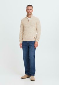 Mann in beigem Half-Zip-Pullover, blauen Jeans und beigen Wildlederschuhen, der vor einem weißen Hintergrund steht.