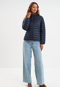 Chaqueta de plumas navy con diseño acolchado horizontal, cierre frontal y cuello alto, combinada con vaqueros de pierna ancha azul claro y Botines marrones.