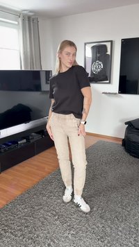 Noor naine mustas t-särgis, beežides pükstes ja valgetes tossudes, seisab hallil vaibal moodsa elutoa keskel, kus on televiisor ja raamitud kunst.