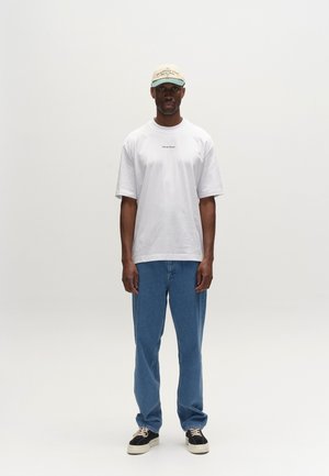 Muž stojaci, má na hlave béžovú čiapku, biele nadrozmerné tričko s malým textom, modré džínsy a čierne tenisky s bielou podrážkou.