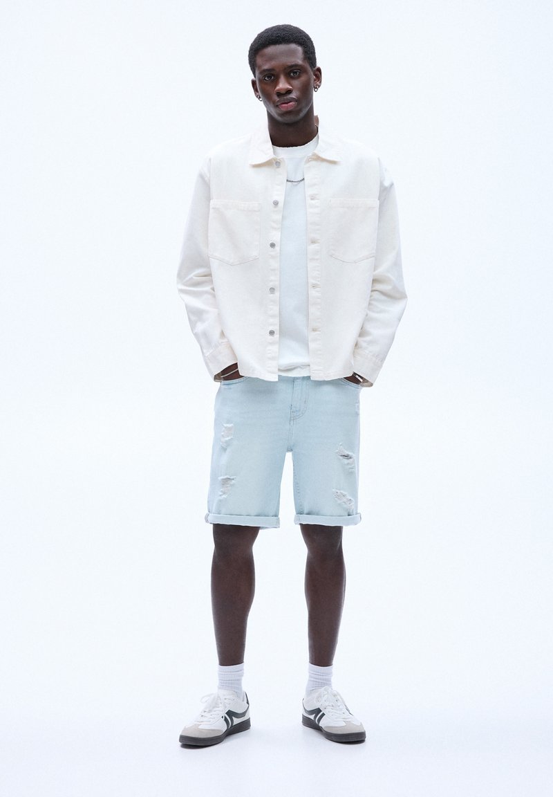 Ung man som står med händerna i fickorna, iförd vit skjorta, krämfärgad jacka, ljusa slitna denimshorts, vita strumpor och sneakers.