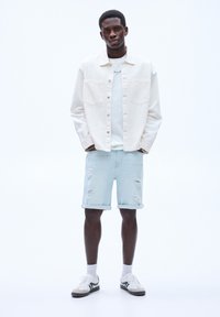 Lysfarvet denimjakke over en hvid t-shirt, parret med lyseblå ripped shorts og hvide sneakers med grønne accenter.