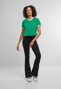 Zielony skrócony T-shirt z białymi wykończeniami w połączeniu z czarnymi spodniami w chmurze. Model nosi białe trampki na gładkim, jasnoszarym tle.