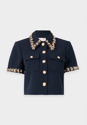 Femme portant une veste courte bleu marine avec bordure florale, un top beige, une jupe plissée bleu marine avec ceinture marron, et des sandales beige à talons hauts, debout.