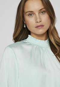 Ljusgrön satinblus med hög smockad krage, draperade ärmar och mjuk lyster. Har en enkel, elegant design.