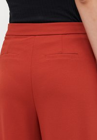 Röda strukturerade byxor med mjuk textur, med två bakfickor och en åtsittande midja, visade bakifrån mot en svart bakgrund.