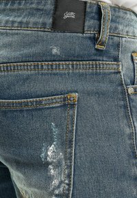 Lys blå denimjeans med slitte områder. Har gule sømmer, et svart lærmerke, og klassisk femlommers design.