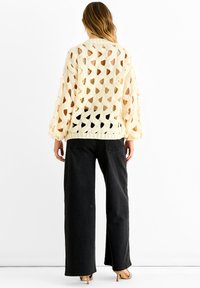 Woman wearing a loose cream knitted sweater with large holes and wide-leg black jeans, standing against a plain white background.