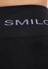 Musta joustava vyötärö, jossa on vaaleanharmaa "SMIL"-teko. Sileä kankaan rakenne, hieman röpelöinen pinta, lähikuva, joka näyttää materiaalin yksityiskohdat.