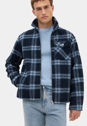 Ung mand iført en marineblå og lyseblå ternet lynlåstrøje over en lyseblå sweater og lyseblå jeans, der står med den ene hånd i lommen.