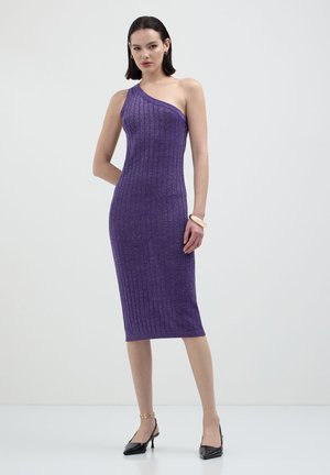 Donna che indossa un vestito midi viola a coste, senza maniche e con una spalla scoperta, abbinato a tacchi a punta neri e gioielli dorati, in piedi su sfondo bianco.