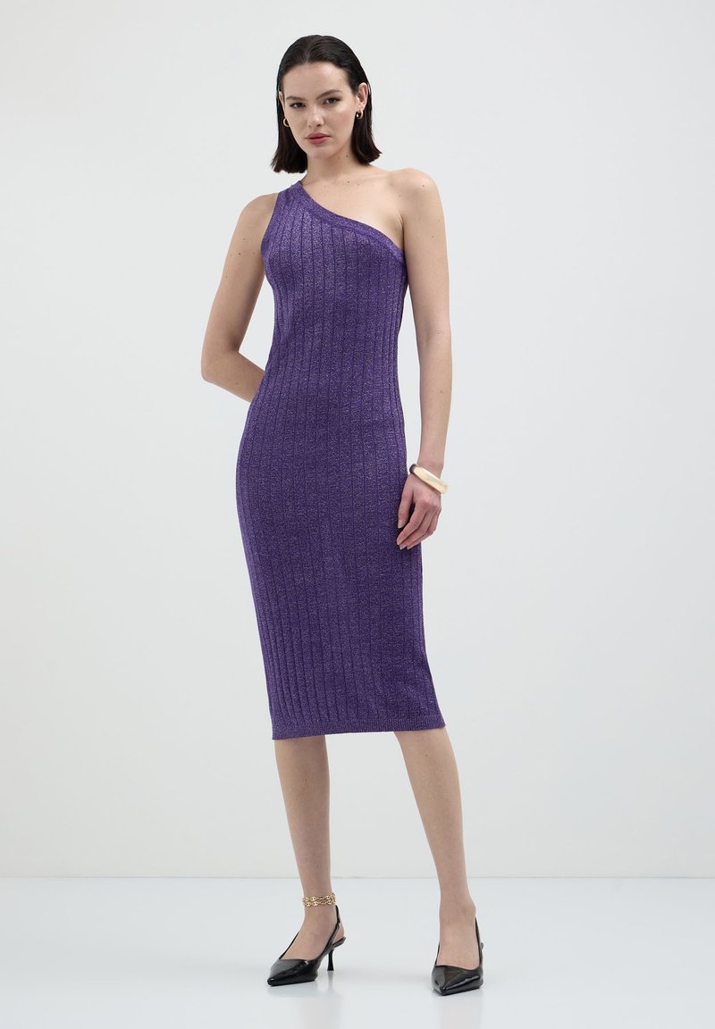 Donna che indossa un vestito midi viola a coste, senza maniche e con una spalla scoperta, abbinato a tacchi a punta neri e gioielli dorati, in piedi su sfondo bianco.