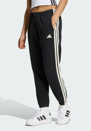 Osoba nosząca czarne spodnie treningowe Adidas z białymi paskami i białe trampki Adidas na jednolitym jasnym tle.