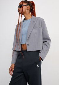 Giovane con lunghe trecce rosse, che indossa occhiali da sole neri, orecchini a cerchio argentati, un blazer grigio corto, un top corto azzurro e pantaloni neri.