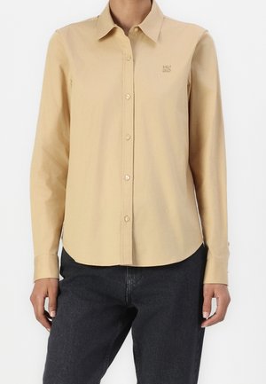 Beige langærmet skjorte med knapper og krave samt lille broderet logo, båret med sorte cowboybukser.