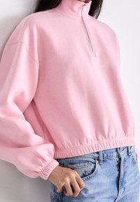 Felpa corta rosa con collo alto, chiusura con zip e orlo elasticizzato. Presenta maniche ampie e un tessuto morbido e strutturato.