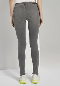 Šedé skinny džíny s hladkou texturou, pěti kapsami a středním pasem, doplněné bílými teniskami s neonově žlutými akcenty.