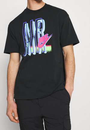 Czarna koszulka z krótkim rękawem z dużym kolorowym napisem „AIR” i różowym logo Nike swoosh na klatce piersiowej, noszona przez osobę z jedną ręką w kieszeni.