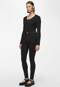 Svart ribbet langermet topp kombinert med høyhåndsydd svart skinny jeans og svarte Mary Jane-sko med chunky hæl. Nøytral bakgrunn.