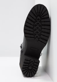 Semelle de botte noire orientée vers l’avant avec un motif profond de bande de roulement, appuyée contre un mur et un sol blancs.