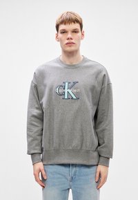 Mladý muž v šedé mikině Calvin Klein a světle modrých džínách, stojící před obyčejným bílým pozadím.