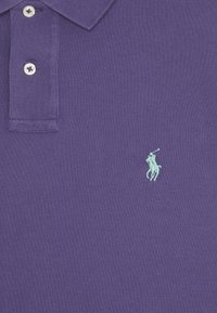 Lila polo skjorta i texturerad bomull, med en tvåknappsslutning och en turkos broderad logotyp av en polospelare.