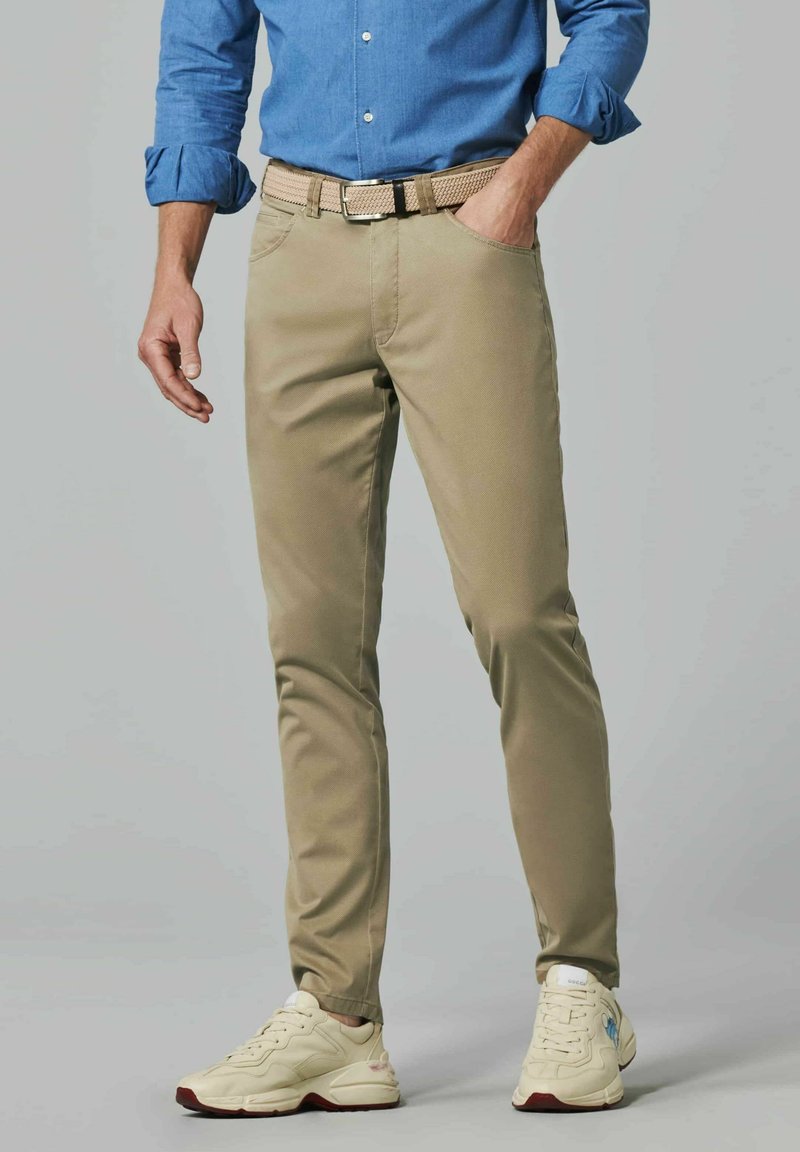 Beige broek in een slim fit, gemaakt van duurzaam materiaal. Heeft een gevlochten riem en is gecombineerd met lichtgekleurde sneakers.