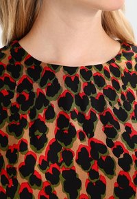 Tissu à motifs floraux dans des tons de rouge, vert et noir sur fond beige ; présente un col rond et une texture lisse.