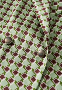 Tissu vert avec un motif géométrique répétitif de carrés marron et blancs, avec deux gros boutons métalliques attachés.