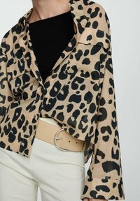 Chemise à imprimé léopard en beige et noir, en tissu léger, coupe courte, manches longues, dotée de deux poches poitrine et d'une fermeture à boutons.