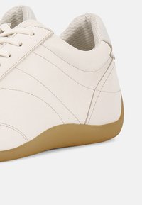 Αθλητικό παπούτσι από λευκό δέρμα με υφή, με καμπύλες ραφές και ανοιχτό καφέ ελαστική σόλα.