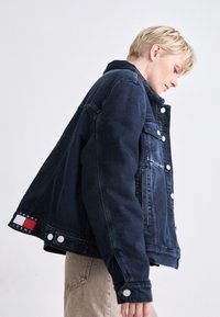 Tamna traper jakna klasičnog dizajna, sa srebrnim dugmadi, džepom na prsima i Tommy Jeans logom na rubu. Glatka tekstura.