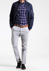 Cazadora bomber azul marino sobre una camisa de cuadros azul y rojo, pantalones chinos gris claro y zapatos negros. La chaqueta cuenta con un bolsillo con cremallera.