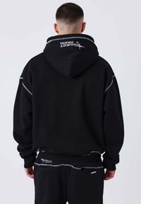 Sudadera negra con una capucha grande, con costuras blancas en contraste y un logo en la parte trasera. Tejido suave con un corte relajado.