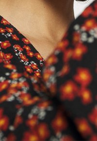 Tessuto nero con un motivo floreale che presenta fiori rossi e gialli e piccoli accenti bianchi. Ingrandimento che mostra la texture e i dettagli del collo.