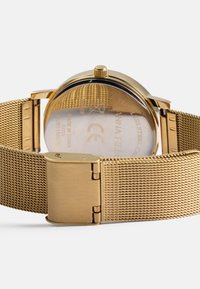 Χρυσό ρολόι από ανοξείδωτο ατσάλι με πλέκτο λουρί, διαθέτει κυκλικό καντράν, διάφανο καπάκι και κούμπωμα, με εγχάρακτη την επωνυμία στην πίσω πλευρά.