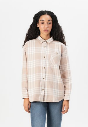 Ung kvinne med krøllete hår iført en beige og hvit rutete skjorte med brystlomme og blå jeans mot en hvit bakgrunn.