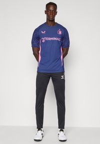 Moški stoji z rokami za hrbtom, oblečen v modro-roza športni dres, črne športne hlače in bele superge na enobarvnem ozadju.